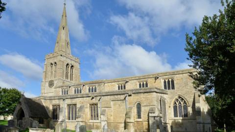 A stone coloured church with a spire on the left hand side. There is a tree on the right and wispy white clouds in the sky.