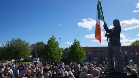 A crowd gathered around a statue of Bobby Sands, with an Irish flag hanging on it. The sky is clear and blue above. There are some trees and a row of houses in the background. 