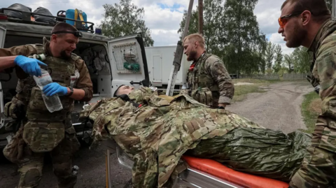 A injured man lies on a stretcher covered with a camouflage jacket is carried to a vehicle with three soldiers taking care of him