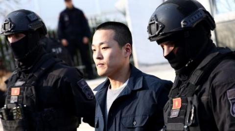 A man being led away by two armed guards wearing heavy duty armour, bulletproof vests, helmets and balaclavas.