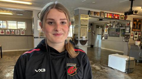 A young lady with blonde hair tied back in a ponytail is looking at the camera. She's wearing a black hoodie with the Dragons rugby team logo on it. In the background is room that has been affected by floodwater. There's a white fridge on the floor to the right of frame and there are many different types of photo frames and shields on the walls. 