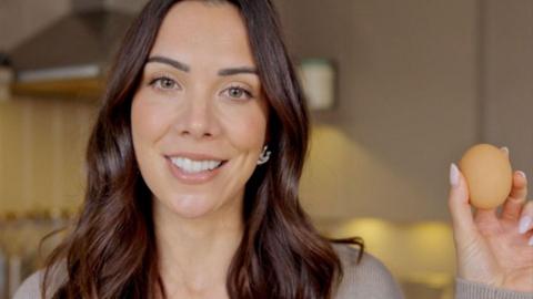 A close-up of a woman’s face with dark long hair. She is smiling and holding a boiled egg up to the camera. She is in a kitchen.