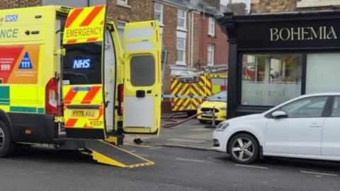 The image shows an emergency response scene on a residential street.
A shopfront with the sign BOHEMIA is visible on the right side. A bright yellow NHS ambulance is parked on the street, with the rear doors open.