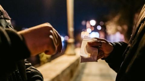 Close up shot of a bag of a powder in a small clear bag being passed to a man on the street. It appears to be dark and there are some lights in the distance.