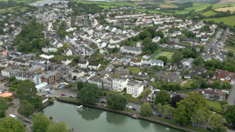 The picture shows an aerial view of a small town surrounded by rolling green hills and countryside. In the foreground, there is a calm river running horizontally across the bottom of the image, with a few small boats moored along its edge and a line of trees bordering the water. Behind the river, there are rows of houses and buildings packed closely together, forming a dense residential area. The houses are mostly two-storey, painted in light colours such as white, cream, and pastel shades, with sloping roofs.