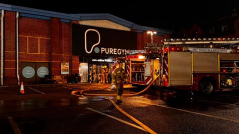 A fire engine is parked with its hose out outside a building with a PureGym sign. There are three fire fighters - one walking away from the building and another two in the doorway.