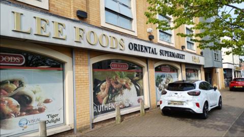 A supermarket called with lettering stating Lee Foods Oriental Supermarket. Pictures of food are on the windows. Some cars are parked nearby. 