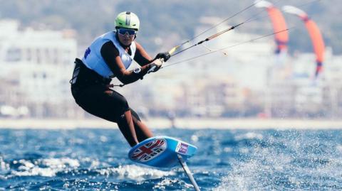 Lily Young is pictured kiteboarding. She is stood on a board on a body of water and gripping a beam that is connected to several wires. She board is airborne and a fin can be seen on its underside. She is facing in the direction of the camera and is wearing a helmet, goggles and a life jacket.