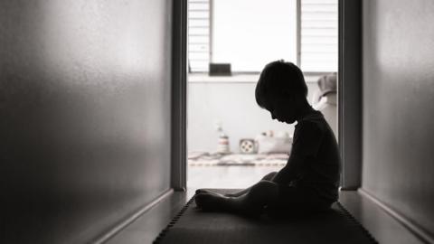 A young child is sitting alone in a hallway in front of an open door.