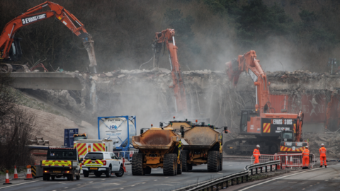 Three large orange cranes demolishing the A432 bridge above the M4. There are various trucks and National Highways vehicles below, as well as several construction workers wearing hi-vis orange overalls and white helmets. There is lots of rubble on the ground and concrete dust in the air.