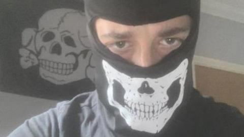 Alfie Coleman wearing a black balaclava with a skull on it. Only his eyes can be seen. Behind him a black flag with a white skull and crossbones is pinned to a wall.