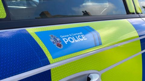 The side of a yellow and blue police vehicle. On the door, above the handle, has the a blue section with the words 'Devon and Cornwall Police'. There is a badge beside it.