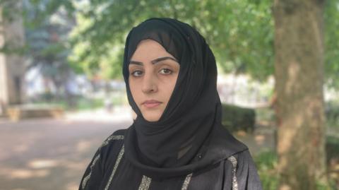 Safia Ahmadei looks directly into the camera wearing a black dress with sparkles and a black hijab. The background is a park