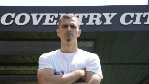 New Coventry City signing Miguel Ángel Brau posed with his arms crossed in front of the stand at the CBS Arena that shows Coventry City on its roof