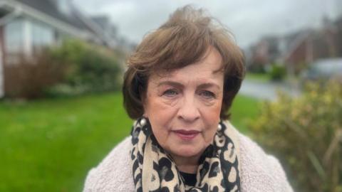 Dodds is looking at the camera. She has short curly brown hair. She is wearing a cream and black patterned scraf and a white coat.