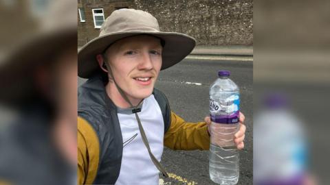 A man wearing a hat and a white and gold top running. He is holding a large water bottle and looking directly at the camera. 