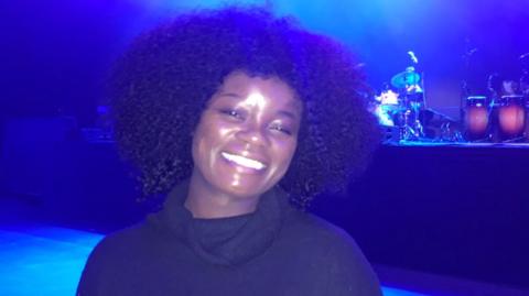 Winnie Ama standing in front of the stage in the Telegraph building in Belfast.  She is smiling at the camera and wearing a black, turtleneck dress.