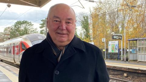 Dennis Fancett is a middle-aged man wearing a blue coat with a scarf tucked inside. He is standing at a railway station with an LNER train just coming in behind him.