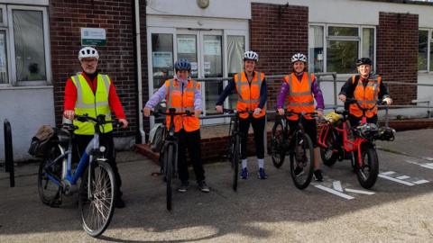 Five people with bikes. The man on the left is wearing an orange zip up jumper, a green high visibility vest and black trousers. The people to the left of him are wearing leggings and zip up jumpers and orange high visibility vests. Behind them is a building made of brick, with a ramp going up to the door. 