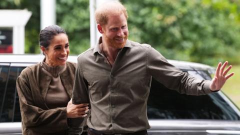 Harry and Meghan wave and smile in Australia. They are wearing khaki green casual outfits and are pictured outside near a car