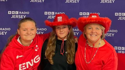 Alice, Maisie and Julia are sitting on a red couch in front of a backdrop with the BBC Radio Jersey logo repeated multiple times. The person on the left is wearing a red hoodie with 'ENG' written on it, the person in the middle is wearing a black hoodie and a red cowboy hat that says 'RED NOSE,' and the person on the right is wearing a red shirt and a large red cowboy hat. All three are smiling at the camera.