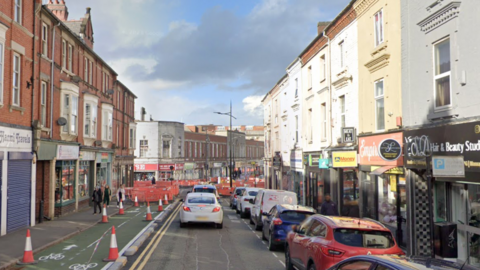 A street view image of a one way street. There are cars parked on the right hand side of the road and on the left there is a cycling lane that is covered in traffic cones, further up there are roadworks visible