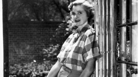 Daphne du Maurier with curled dark hair, wearing a check shirt and trousers, leaning against a doorframe