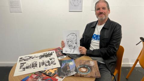 A man in a black leather jacket with a white t-shirt underneath holds up a black and white sketch of a zombie's head. He is sat at a table which is covered in comic books and art