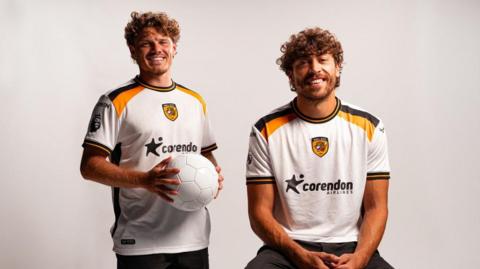 Two men model the Hull City away kit with one of them holding a football. The shirt is white with black and amber stripes round the end of the sleeves and along the shoulder. The club Tiger badge in the middle with the corendon airlines sponsor underneath.