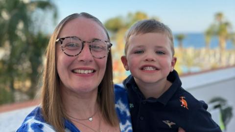 Jennifer Taylor, holding her son William. Jennifer is wearing a blue-and-white floral dress and has long brown hair. She is wearing tortoiseshell glasses. William is in a navy shirt with dinosaurs embroidered on it.