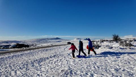 Three people in snow gear walking on snow covered fields under a blue sky.