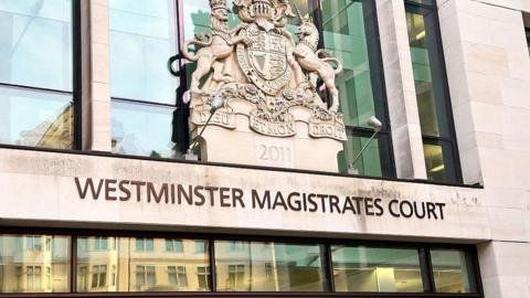 The outside of Westminster Magistrates Court. It has glass windows which have a reflection of buildings in them. And the sign above the windows says 'Westminster Magistrates Court', which is below a historical crest. 