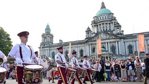 Four members of a drum band walking.
The are wearing white shirts. burgundy trousers and burgundy hats.
There are people watching from the pavement beyond.