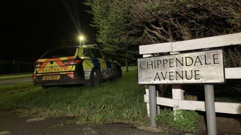 A white sign with the words Chippendale Avenue on it in black. A marked police car is in the background - it has blue and yellow colouring.