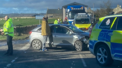 A crash scene with a grey car in the centre badly damaged and a number of emergency vehicles surrounding it 