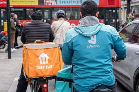 Deliveroo and Just Eat takeaway delivery cycle couriers on a main road, near a pedestrian crossing with traffic lights. The Deliveroo driver is on the right. There is a car to the right of the Deliveroo driver and a TfL bus can be seen ahead. There are pedestrians in the distance.
