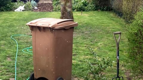 A garden with a brown bin situated slightly off centre to the left, with a hose, tree and a fork pictured across the garden. 