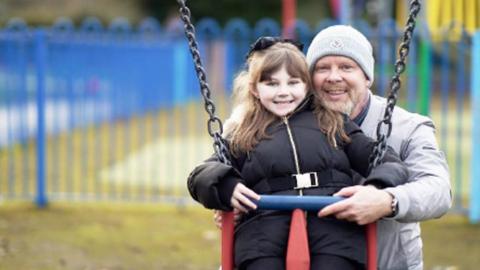 John Stephenson with his granddaughter. She is sitting on a swing and smiling into the camera, as is Mr Stephenson. He is wearing a grey jacket and grey woolly hat.