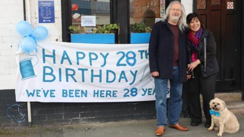 Chantana Parkinson stands outside her shop alongside councillor Phil Riley. A white banner with blue writing includes an image of a sewing needle and cotton reel and the words 'Happy 28th birthday. We've been here 28 years'. Blue balloons are tied to the shopfront and Ms Parkinson's small cream-coloured dog is wearing a matching blue bow tie.