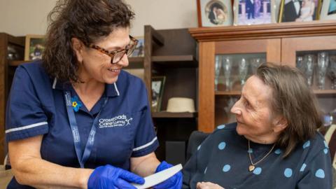 A woman with glasses in a Crossroads Care Surrey uniform talks to an elderly woman in the spotted jumper 