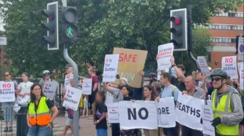 Protestors fill a road, standing on a pedestrian crossing while the traffic lights are red. They are holding up white signs that read "no more deaths" and "B is for Better". A tree and a multi-storey building can be seen in the background.