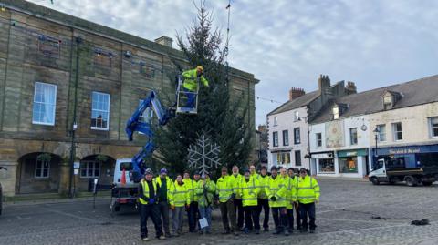 A Christmas tree in Alnwick's main square with a dozen volunteers wearing yellow hi-vis jackets standing in front of it