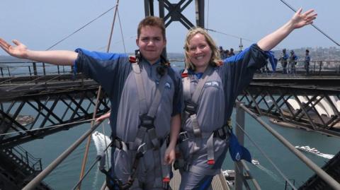 Connor Calleja has short brown hair and has his hand outstretched while wearing harness equipment next to a woman with short blonde hair, also wearing harness equipment, with her hand outstretched to the other side. They appear to be standing on a bridge and water can be seen far below with some boats.