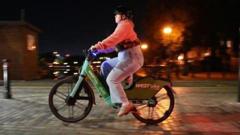A woman in blue jeans, pink sweatshirts and black helmet rides a Dott and West Bike branded e-bike along a cobbled road at night time.  