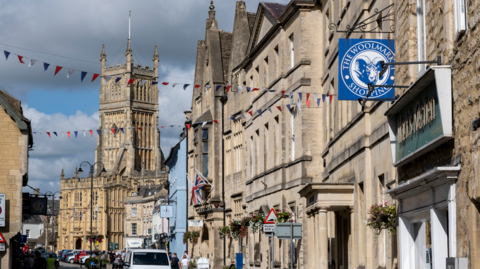 A street view of Cirencester. There are stone buildings with bunting hanging across the street. There is a church in the background.