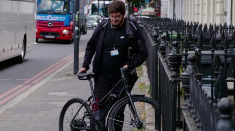 Bilal, dressed in black, holds a bike next to a road and black spikey railings