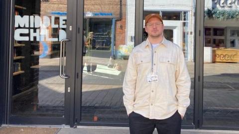 A man wearing glasses, a cream shirt and brown cap is standing in front of a theatre front, made up of large window panels, with a sign reading "Middle Child" in white, cartoon-like letters on the left.