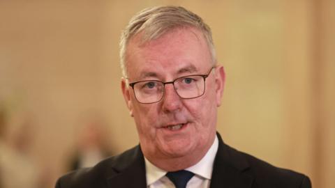 Mike Nesbitt who has short grey/white hair and is wearing thin black framed glasses. He is mid sentence answering a question in Stormont. He is also wearing a black suit and tie with a white shirt. 