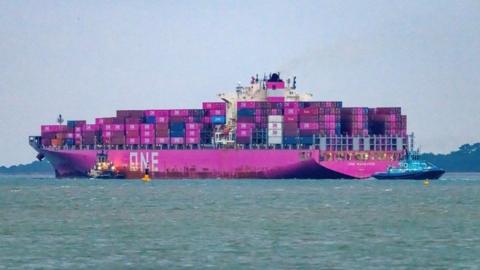 Bright pink ship loaded with a large number of pink containers and some dark blue under sail with two tugs seen behind and to the side