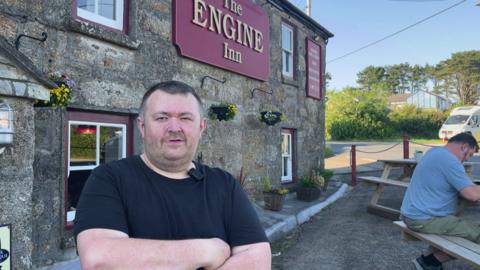 A man in a black t shirt looks at the camera , he runs a pub called The engine Inn , the granite pub exterior is behind him. 